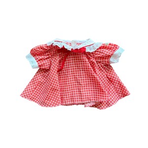 May include: A short-sleeved doll dress featuring a red and white grid pattern, a white eyelet lace collar, and white eyelet lace trim on the sleeves. A thin red ribbon is tied in a bow at the center of the neckline, adding a decorative detail.