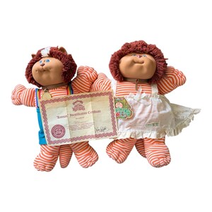 May include: Two plush Koosas dolls with brown yarn hair and orange-and-white striped bodies. One doll wears blue overalls with rainbow straps and a gold medallion, while the other wears a white apron over its striped body. A paper certificate is included.