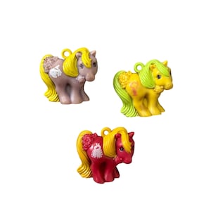 May include: Three small plastic pony figurines with molded manes and tails, each featuring a loop on the back for hanging. One is light purple with yellow hair, one is yellow with light green hair, and one is red with yellow hair and floral details.
