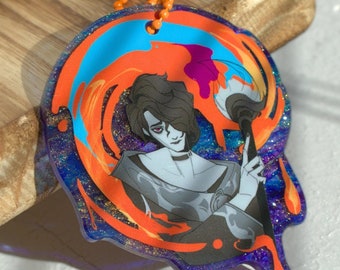 May include: A round, clear acrylic keychain featuring a stylized illustration of a person holding a paintbrush. The design incorporates vibrant orange, blue, and purple paint strokes. The keychain has an orange ball chain for attachment.