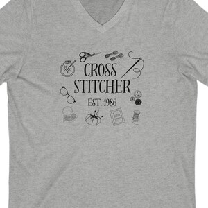 May include: A grey V-neck t-shirt with the text "CROSS STITCHER EST. 1986" and illustrations of sewing tools. The design includes scissors, needles, buttons, and other craft-related icons.
