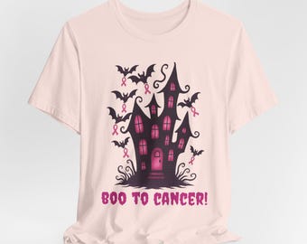 以下が含まれることがあります： ピンク色のTシャツ。濃い紫色の幽霊屋敷のデザイン、ピンクのアクセント、ピンクのリボンをつけたコウモリが特徴です。「BOO TO CANCER!」の文字がピンク色でプリントされています。乳がん啓発のデザインです。