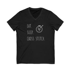 May include: Black V-neck t-shirt with white text and a cross-stitch design. The text reads "EAT. SLEEP. CROSS STITCH." A white illustration of a cross-stitch hoop and needle is also present.
