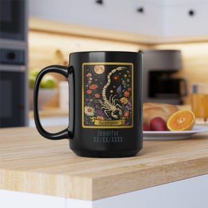 May include: Black ceramic mug featuring a detailed illustration of a scorpion, flowers, and a full moon. The mug has the word "Scorpio" and the name "Jennifer" printed on it. A large, black handle is on the left side.