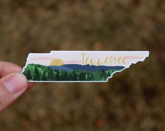 May include: A sticker of the state of Tennessee with a sunset scene over a mountain range. The sticker is white with a pink sky, green trees, and blue mountains. The word "Tennessee" is written in gold lettering.