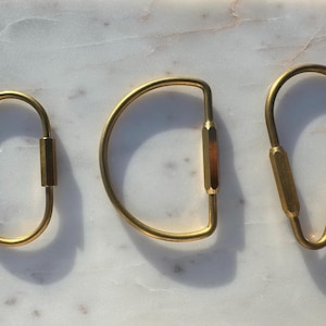 May include: Three gold-toned keychains with different shapes. The keychains are made of metal and have a simple, minimalist design.