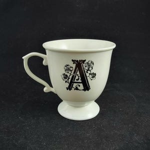 Może przedstawiać: Kremowy ceramiczny kubek z czarną literą "A" i kwiatowym wzorem. Kubek ma zakrzywiony uchwyt i podstawę. Wz&oacute;r jest wyśrodkowany z przodu kubka.