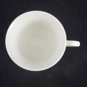Może przedstawiać: Widok z g&oacute;ry na białą ceramiczną filiżankę do herbaty z uchwytem. Filiżanka jest pusta i stoi na ciemnej powierzchni. Wnętrze filiżanki jest gładkie, a uchwyt ma prosty kształt.
