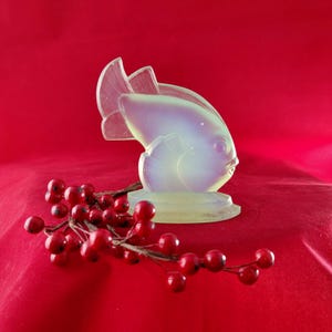 Puede incluir: Una escultura de pez de cristal translúcido con reflejos iridiscentes, sobre un fondo rojo. El pez tiene aletas detalladas y una base redonda. Una ramita de bayas rojas añade un elemento de color contrastante.