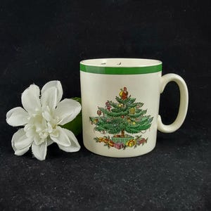Puede incluir: Una taza de cerámica blanca con un borde verde y un diseño de árbol de Navidad. El árbol está decorado con adornos, regalos y una estrella en la parte superior.