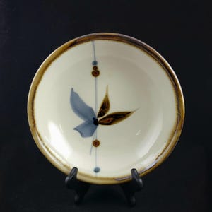 Könnte beinhalten: Eine weiße Keramikschüssel mit braunem Rand und einem blauen und braunen Blumendesign. Die Schüssel steht auf einem schwarzen Ständer.