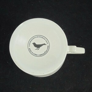 Może przedstawiać: Biały ceramiczny kubek z uchwytem, ​​widziany od dołu. Kubek posiada czarną sylwetkę ptaka w okrągłym wzorze z napisem "Dishwasher & Microwave Safe Made in China Fabriqu&eacute; en Chine."