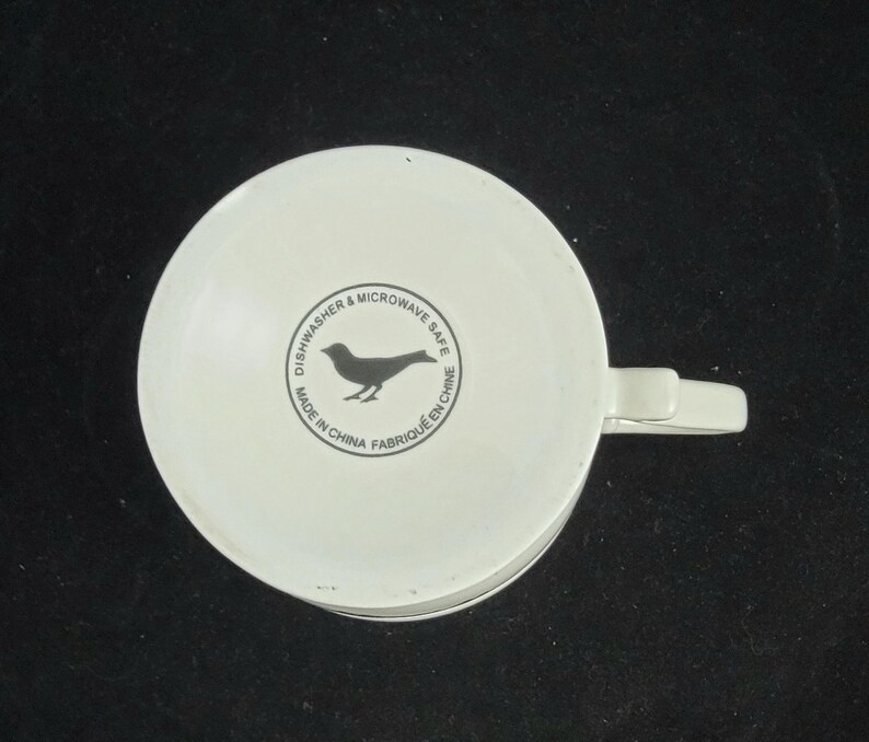 Może przedstawiać: Biały ceramiczny kubek z uchwytem, ​​widziany od dołu. Kubek posiada czarną sylwetkę ptaka w okrągłym wzorze z napisem "Dishwasher & Microwave Safe Made in China Fabriqu&eacute; en Chine."