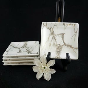 Może przedstawiać: Zestaw białych kwadratowych talerzy z marmurowym wzorem. Talerze są ułożone w stos i prezentowane na czarnym stojaku. Biały kwiat na pierwszym planie. Idealne do serwowania przystawek lub deserów.