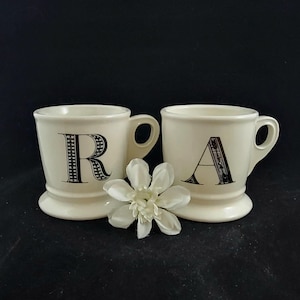 Può includere: Due tazze in ceramica bianca con disegni di lettere nere. La tazza a sinistra ha la lettera "R" e la tazza a destra ha la lettera "A".