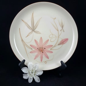 Puede incluir: Plato de cerámica color crema con diseño floral. El plato presenta una gran flor rosa en el centro, con hojas grises y tallos marrones. El borde es marrón oscuro. Una flor blanca está en primer plano.