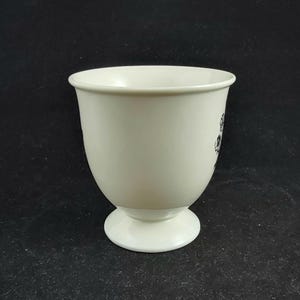 Może przedstawiać: Kremowy ceramiczny kubek z podstawą. Kubek ma zaokrąglony korpus i szeroki otw&oacute;r. Czarny kwiatowy wz&oacute;r jest widoczny z boku. Kubek jest ustawiony na ciemnym tle.