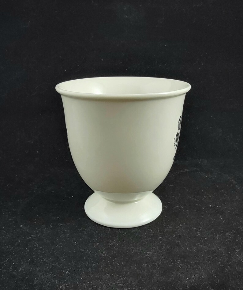 Może przedstawiać: Kremowy ceramiczny kubek z podstawą. Kubek ma zaokrąglony korpus i szeroki otw&oacute;r. Czarny kwiatowy wz&oacute;r jest widoczny z boku. Kubek jest ustawiony na ciemnym tle.