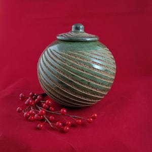 Puede incluir: Un jarrón de cerámica verde con tapa, con líneas horizontales talladas. El jarrón está sobre un fondo rojo, con una ramita de bayas rojas en primer plano. El diseño del jarrón es a la vez decorativo y funcional.