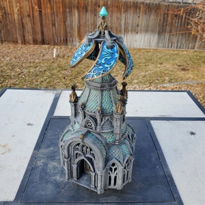 May include: A detailed miniature model of a gothic tower with a blue and gold spire. The tower has a green roof and intricate stonework.