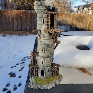 May include: A miniature stone tower with wooden scaffolding and a ladder. The tower is weathered and has a small opening at the base. The tower is set on a base of brown rock with green moss.