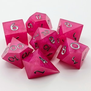 May include: A set of seven polyhedral dice in shades of pink. Each die has silver numbering. The dice are designed for tabletop role-playing games. The dice are made of a translucent material.