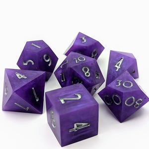 Könnte beinhalten: Ein Satz von sieben violetten Würfeln mit silbernen Zahlen. Die Würfel sind aus einem durchsichtigen Material gefertigt und haben eine gesprenkelte Oberfläche. Die Zahlen auf den Würfeln sind klar und leicht zu lesen.