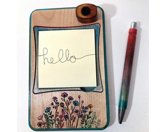 May include: Wooden notepad with a teal border and a floral design. The notepad has a built-in pen holder and a sticky note with the word "hello" written on it. A teal and red pen is next to the notepad.