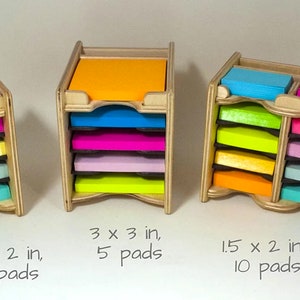 May include: Wooden organizer with five slots for holding sticky note pads. The organizer is made of light wood and has a wavy bottom edge. The sticky note pads are in various colors, including pink, yellow, blue, green, and orange. The text below the organizer reads "1.5 x 2 in, 5 pads" and "3 x 3 in, 5 pads".