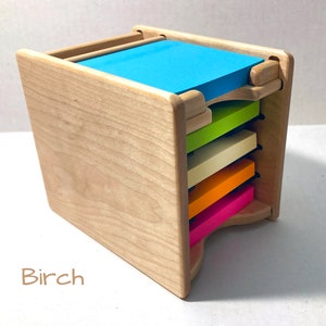 May include: A wooden note pad holder with a stack of colorful sticky notes. The holder is made of birch wood and has a slot for each note pad. The note pads are in the colors pink, orange, yellow, green, and blue.
