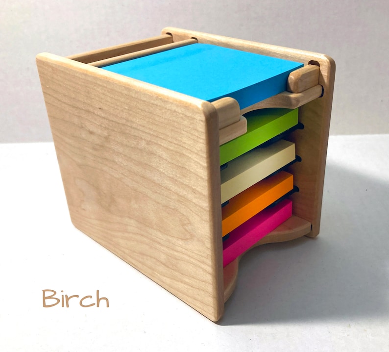 May include: A wooden note pad holder with a stack of colorful sticky notes. The holder is made of birch wood and has a slot for each note pad. The note pads are in the colors pink, orange, yellow, green, and blue.