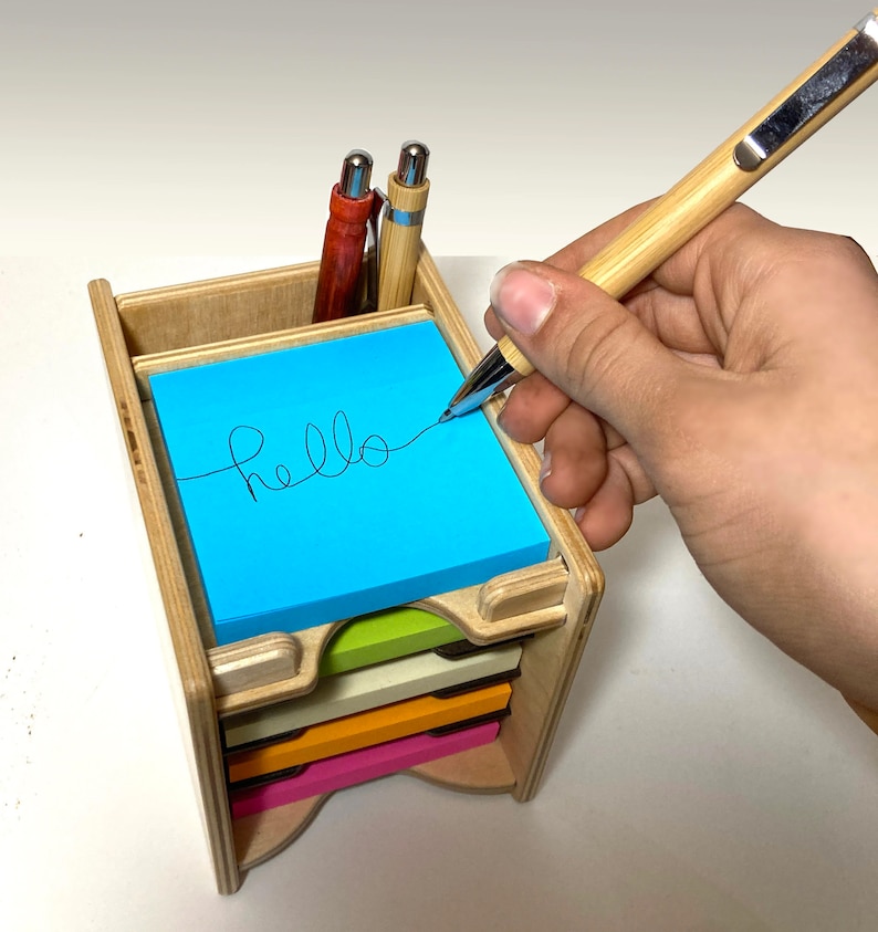 May include: A wooden desk organizer with a stack of colorful sticky notes. The top note is blue and has the word "hello" written on it. A hand is holding a pen and writing on the note.