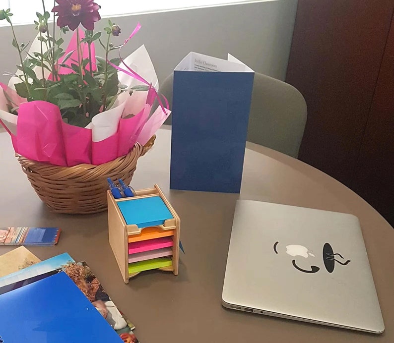 May include: A wooden desk organizer with colorful sticky notes, a blue folder, a laptop with a coffee cup sticker, and a bouquet of pink and purple flowers in a wicker basket.