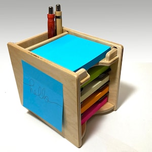 May include: A wooden desk organizer with multiple compartments for storing sticky notes and pens. The organizer is made of light wood and has a natural finish. The top compartment has a blue sticky note with the word "hello" written on it. Two pens are visible in the top compartment.
