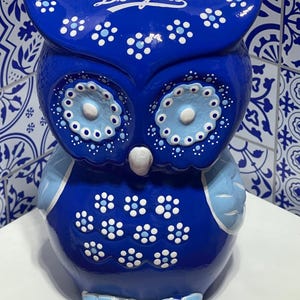 May include: A ceramic owl figurine in shades of blue, adorned with white floral patterns and the word "Dodgers." The owl has large, round eyes and a light blue chest and feet. The background features a blue and white tile pattern.