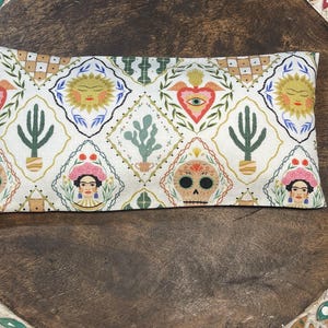 May include: Rectangular cream-colored fabric item with a repeating pattern of suns, hearts, cacti, and sugar skulls. The design includes a Frida Kahlo-inspired portrait. The item is likely a decorative pillow or eye mask.