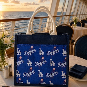 May include: A square-shaped tote bag with a dark blue jute exterior and two white rope handles. The front panel features a repeating pattern of the Los Angeles Dodgers logo and baseball graphics on a bright blue background with a glittery trim.