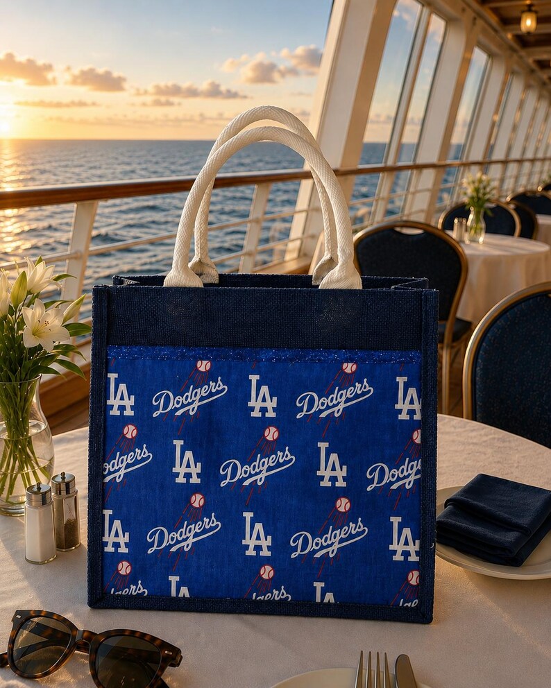 May include: A square-shaped tote bag with a dark blue jute exterior and two white rope handles. The front panel features a repeating pattern of the Los Angeles Dodgers logo and baseball graphics on a bright blue background with a glittery trim.