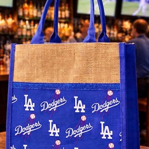 May include: A blue and tan tote bag featuring a repeating pattern of the Los Angeles Dodgers logo, the word Dodgers in script, and small baseball graphics. The bag has two blue fabric handles and a natural burlap upper section with blue trim.