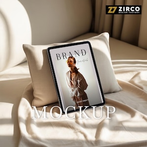 May include: A digital tablet displaying a magazine cover with the word "BRAND" in bold letters. The image features a woman wearing sunglasses. The word "MOCKUP" is displayed at the bottom of the tablet. The tablet rests on a pillow and blanket.