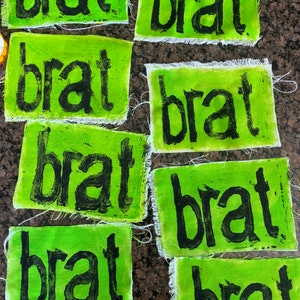 May include: Eight green fabric patches with the word "brat" printed in black paint. The patches have frayed edges.