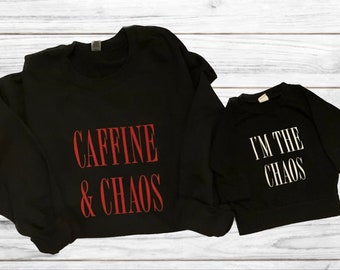 May include: Two black long-sleeved shirts with a matching design. The larger shirt says "CAFFINE & CHAOS" in red and white lettering. The smaller shirt says "I'M THE CHAOS" in white lettering.