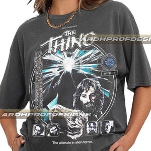 Pode incluir: Camiseta cinza escuro com um gráfico de "The Thing" de John Carpenter. O design inclui o título do filme, uma ilustração central e o texto "The ultimate in alien terror."