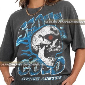 May include: Dark gray t-shirt featuring a distressed graphic of a skull with a red eye and blue flames. The text "STONE COLD" and "STEVE AUSTIN" are printed in blue. The shirt has short sleeves.