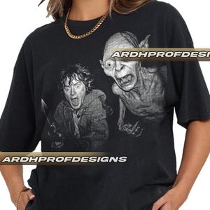May include: Black t-shirt featuring a grayscale image of two characters from The Lord of the Rings. One character is holding a knife and screaming, while the other has a wide-eyed expression. The text "ARDHPROFDESIGNS" is printed on the shirt.
