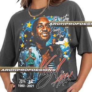 Op de afbeelding: Donkergrijs t-shirt met een portret van een man, sterren en tekst. Het shirt toont de naam "Virgil Abloh" in een gestileerd schrift en de jaren "1980-2021". Het ontwerp heeft een vintage, distressed look met blauwe en gele tinten.