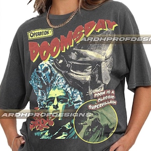 May include: Dark gray t-shirt featuring a graphic design with the text "OPERATION DOOMSDAY" in red and yellow. The design includes illustrations of a masked figure and the words "DOOM IS A CLASSIC SUPERVILLAIN".