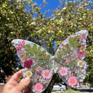May include: Butterfly-shaped resin art piece with embedded flowers and small stones. The wings feature pink, white, and red flowers, along with green foliage, set against a clear background. The piece is held up against a backdrop of green trees and a blue sky.