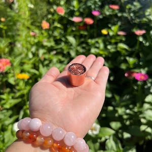May include: A cylindrical copper-colored object with a coiled wire top sits on a palm, accompanied by two beaded bracelets. One bracelet features smooth, pale pink round beads, while the other consists of translucent, deep orange carnelian beads.