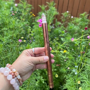 May include: A copper-colored crystal wand with a clear crystal tip, wrapped with copper wire. The wand is held against a backdrop of green foliage and colorful flowers.